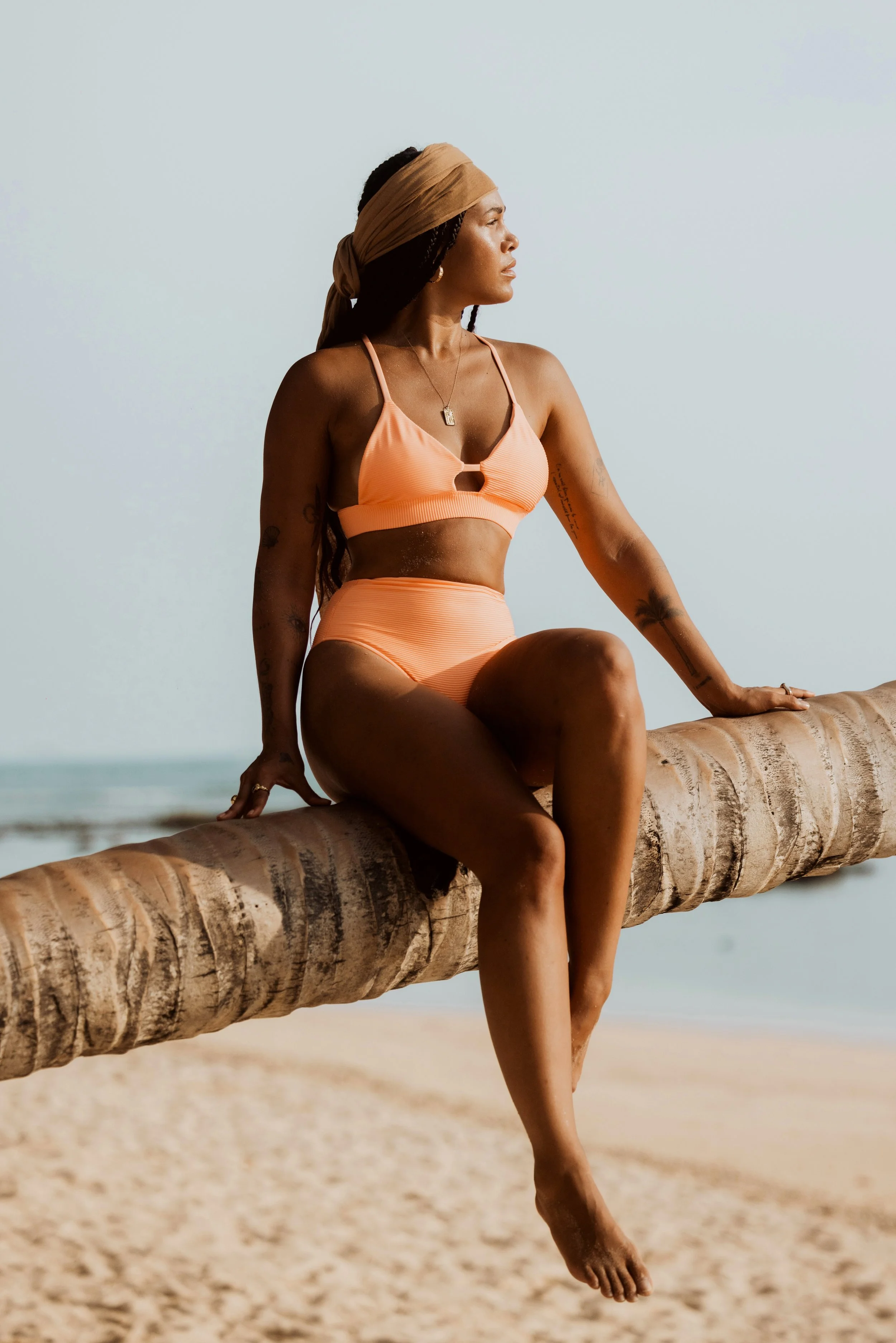 Woman in orange swimsuit sitting on a palm tree trunk at the beach, looking into the distance.