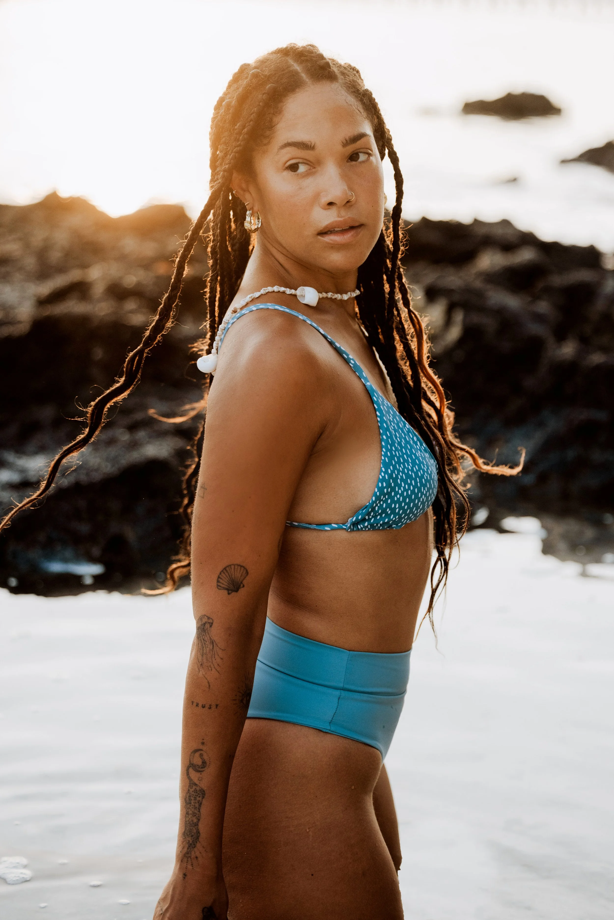 Person with long braided hair wearing a blue bikini, standing on a beach with a rocky background, under natural sunlight.