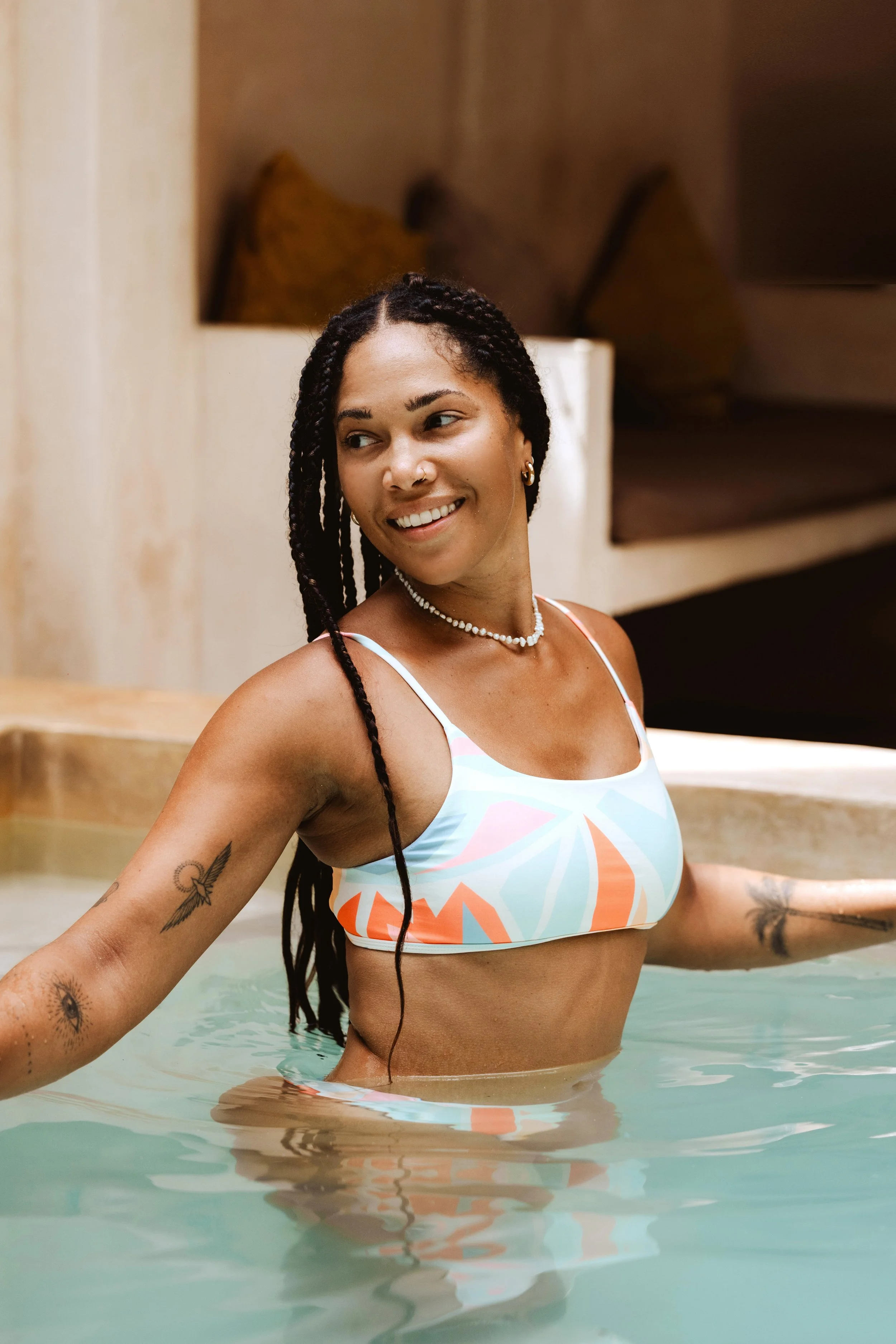 Person with braids in a colorful swimsuit standing in a pool, smiling and showing arm tattoos.