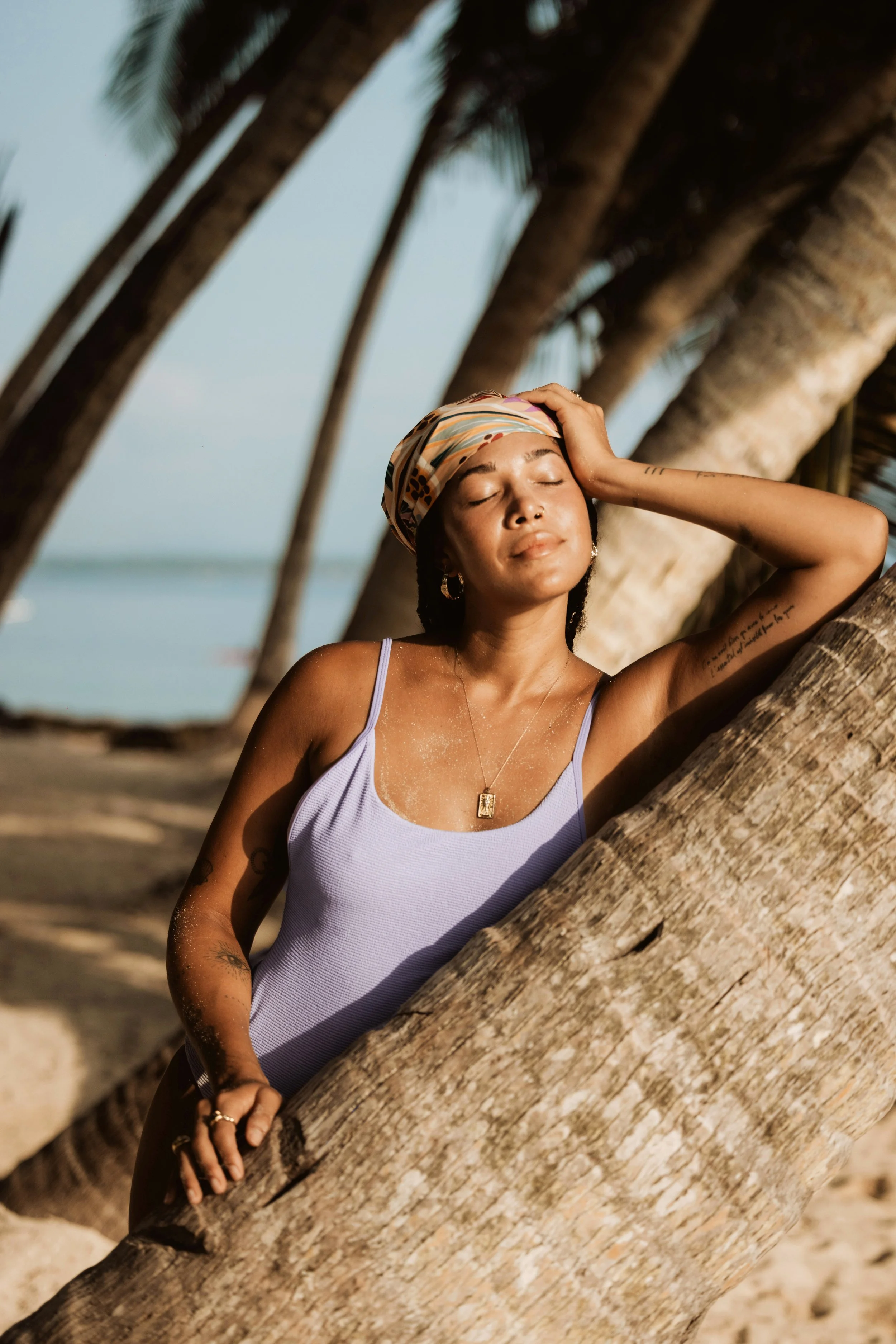 Person wearing a purple swimsuit and headscarf, standing against a palm tree on a beach.