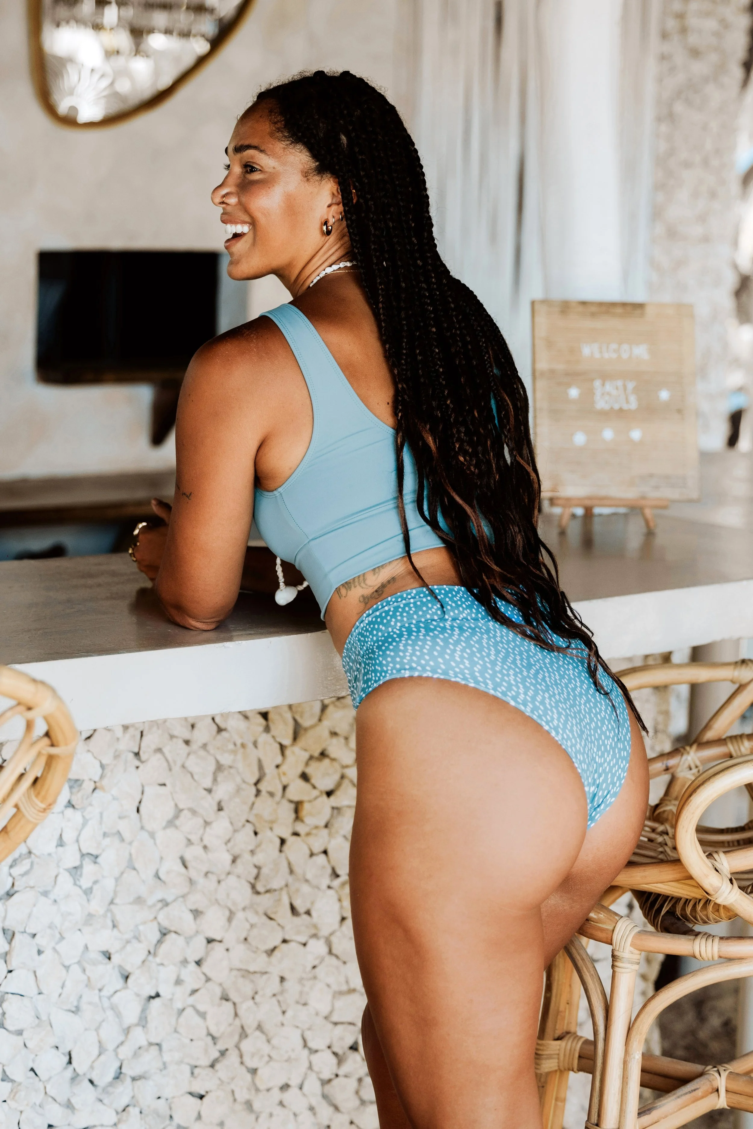 Woman in blue bikini leaning against a bar, smiling, with braided hair.