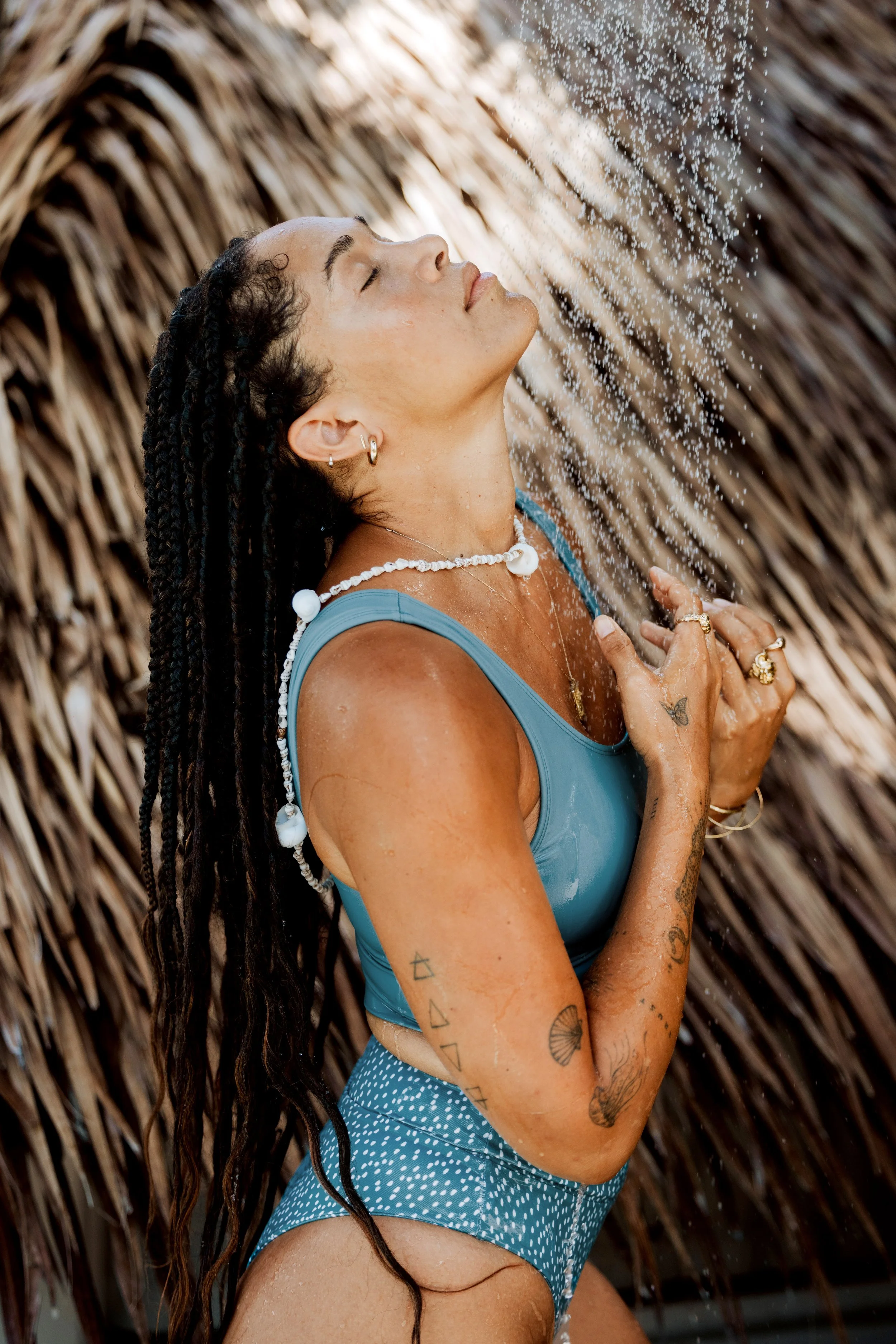 Person with braided hair in a blue swimsuit showering outdoors, surrounded by palm fronds, eyes closed, wearing shell necklace and jewelry, with visible tattoos.