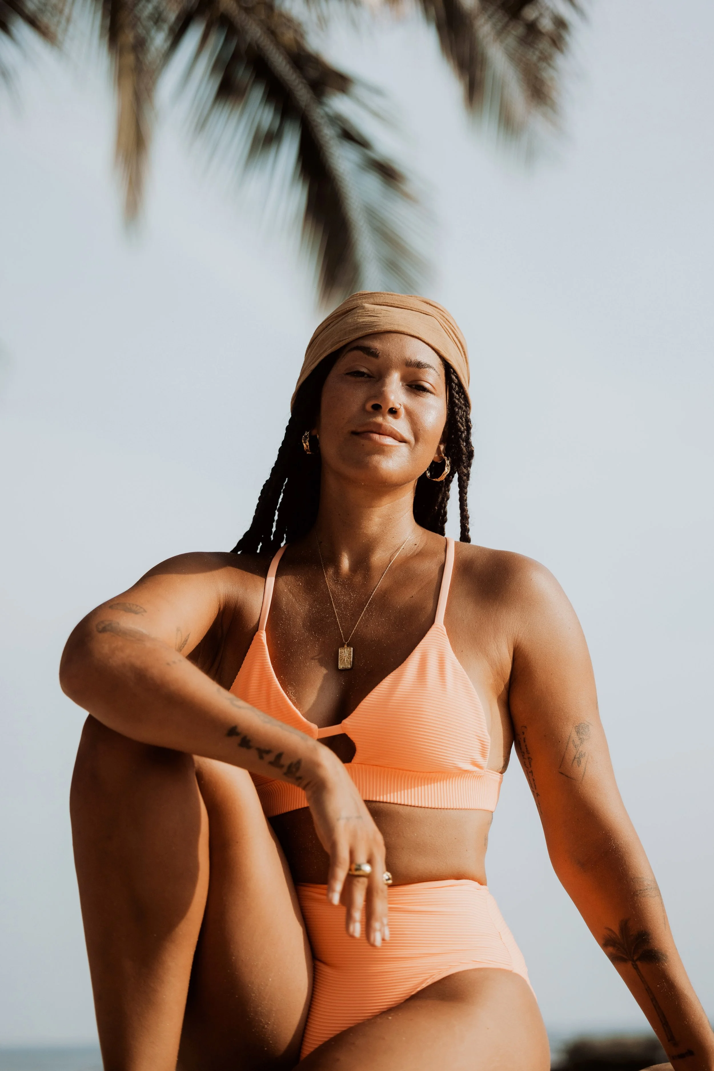 Woman in coral swimsuit sitting outdoors, wearing headscarf and jewelry, with palm tree background.