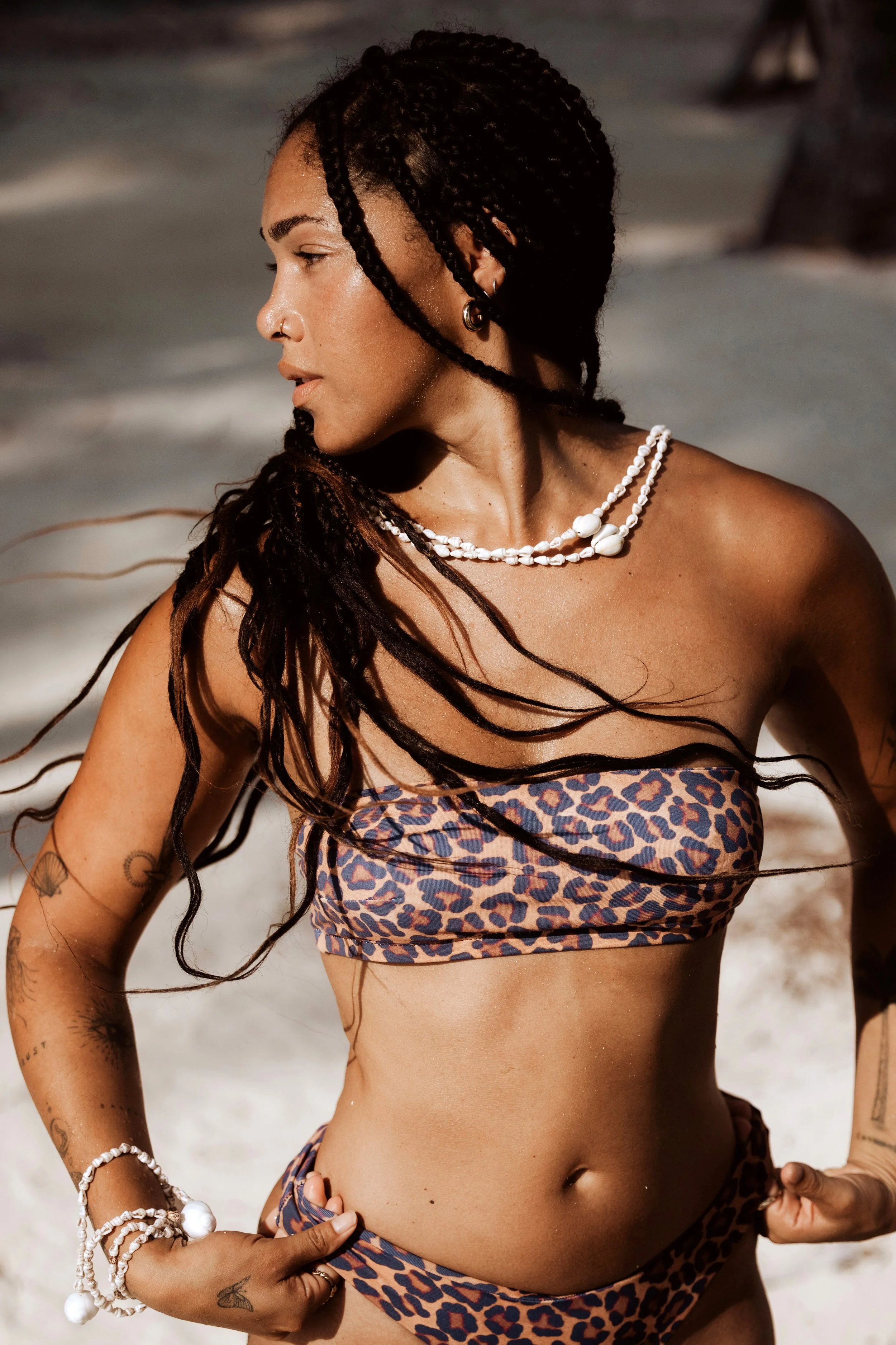 Woman with braided hair wearing a leopard print bikini and shell jewelry on a beach.