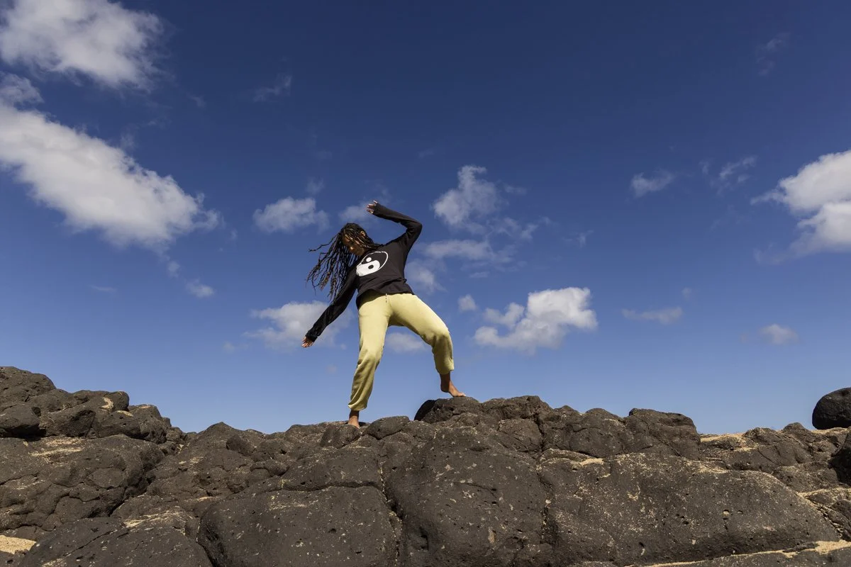 Person with dreadlocks wearing a yin-yang sweater and yellow pants, standing on rocky terrain under a blue sky with clouds.