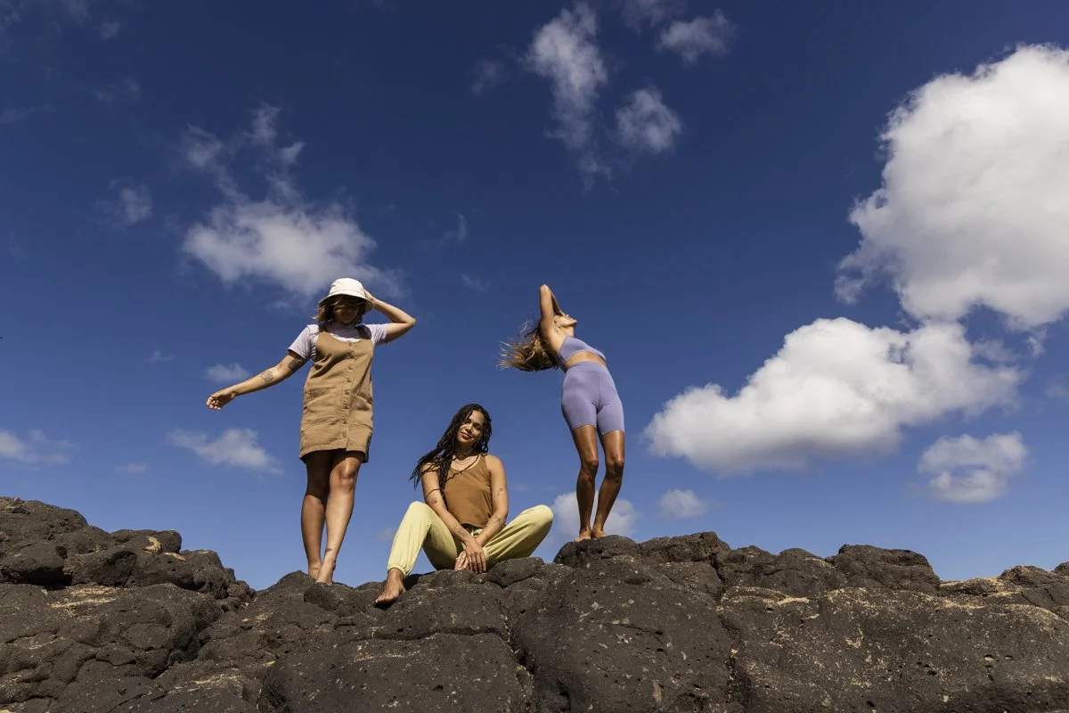Three women standing and sitting on black rocks under a blue sky with clouds. One is wearing a tan dress and bucket hat, another is seated in yellow pants, and the third is raising her arms in a blue outfit.