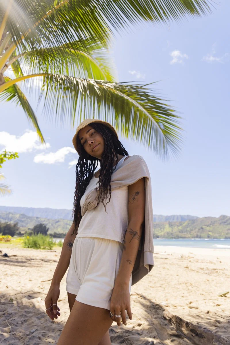 Person standing on a beach under a palm tree, wearing a hat and casual summer clothing, with mountains and ocean in the background.