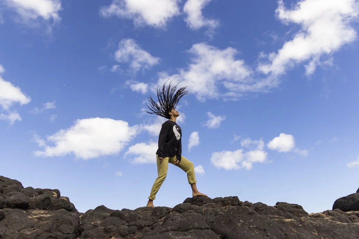 Person with long hair performing a hair flip on rocky terrain against a blue sky with clouds.