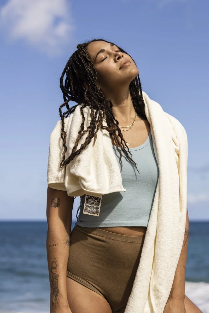 Person with braided hair and tattoos standing by the ocean, wearing a light blue tank top and brown shorts, with a white towel draped over their shoulders.