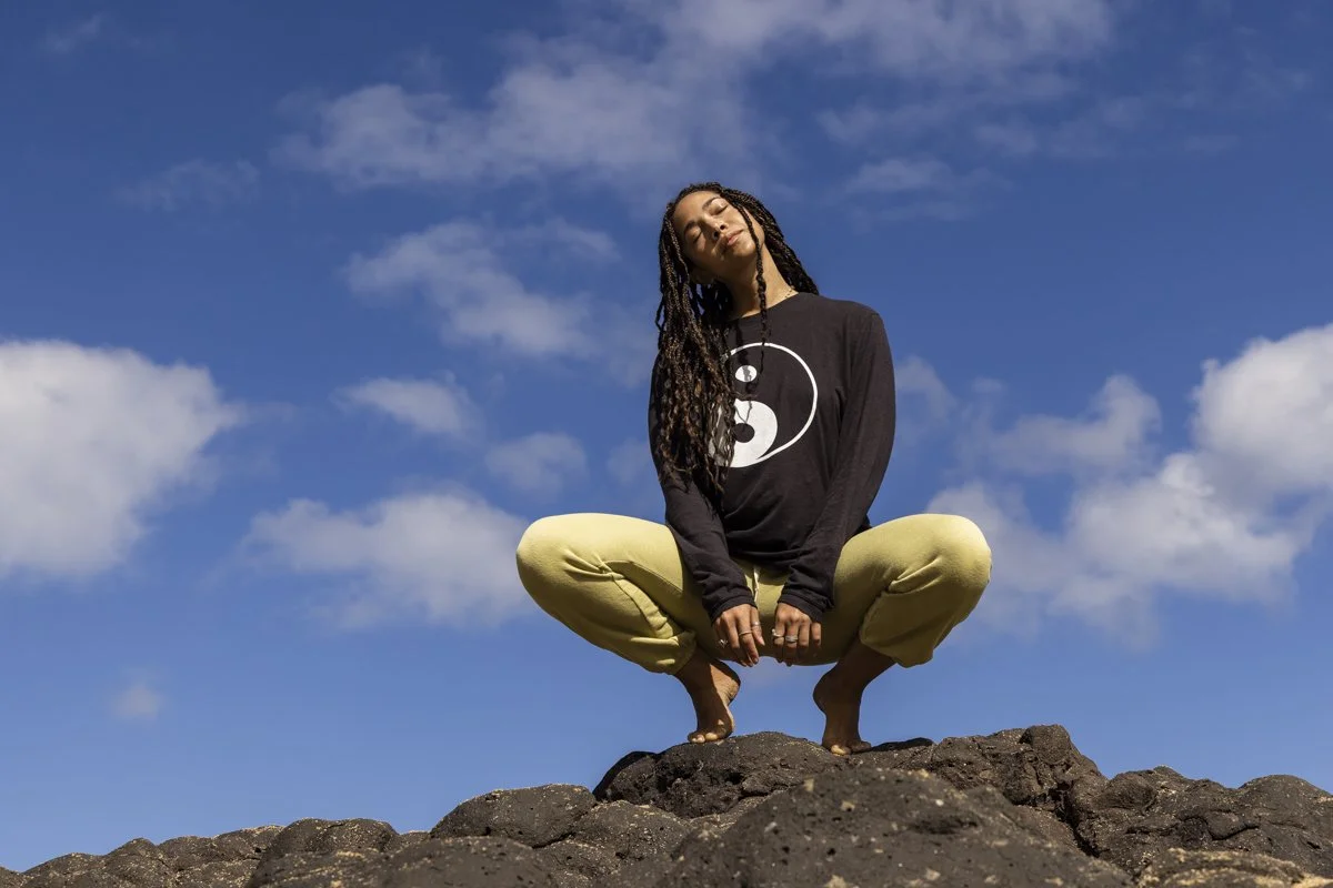 Person squatting on rocks against blue sky, wearing a black yin-yang shirt and yellow pants.