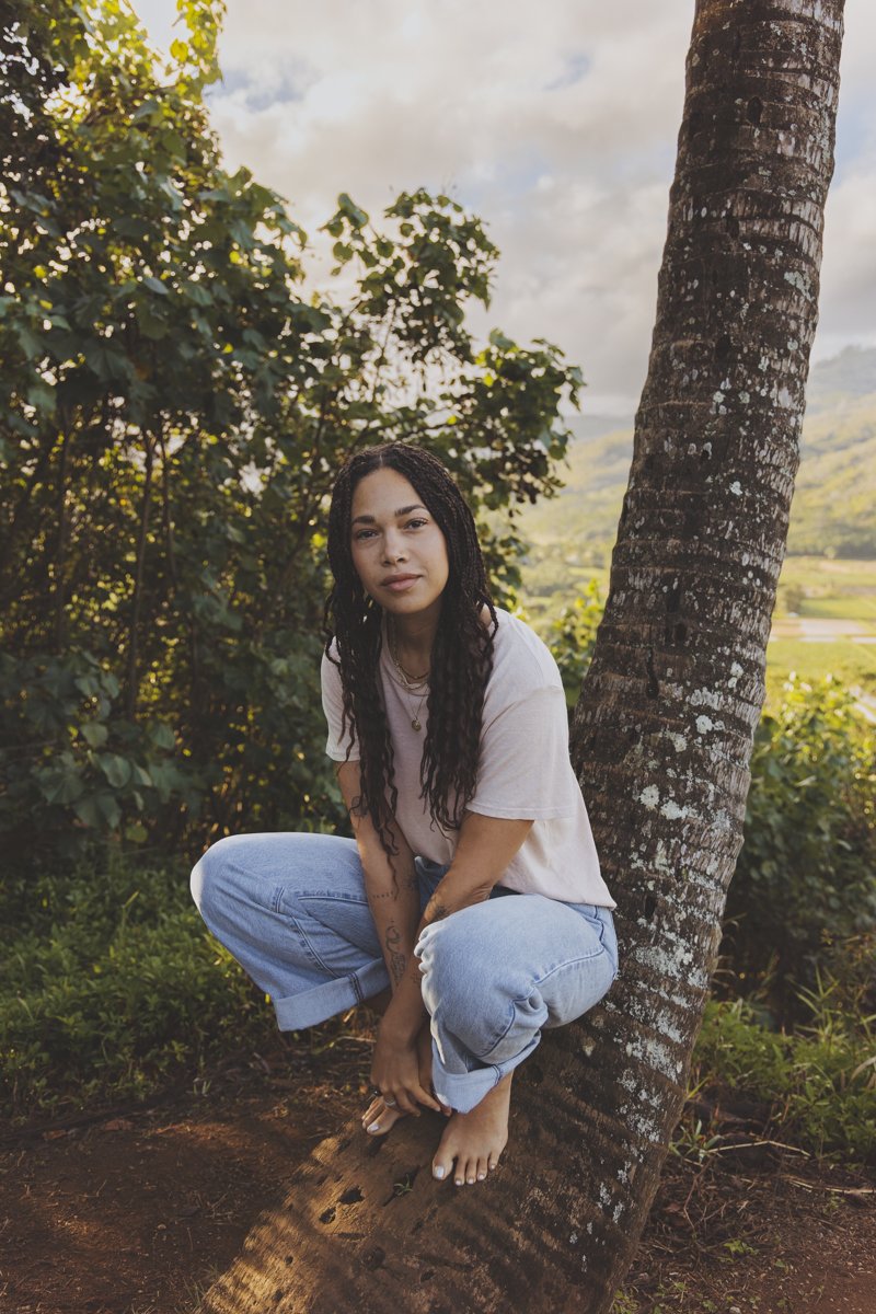 Person with long braided hair crouching on a tree, wearing a casual outfit, in an outdoor nature setting.