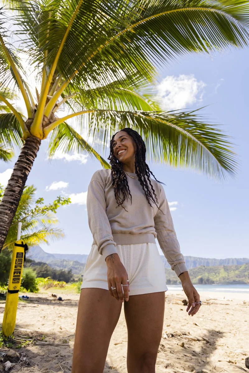 Woman in casual clothing standing on a beach under a palm tree, with a blue sky and mountains in the background.