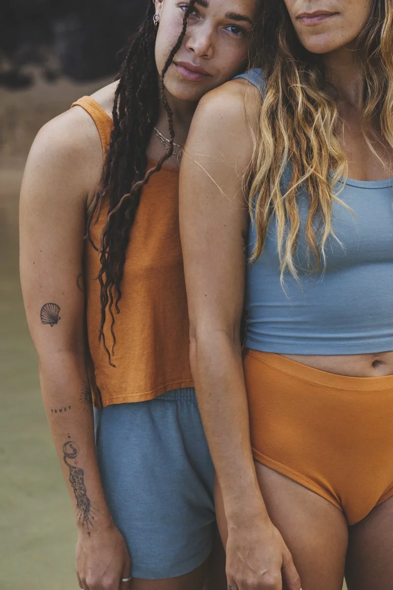 Two women in casual summer outfits, one in orange and blue, the other in blue and orange, standing closely with tattoos visible on arms.
