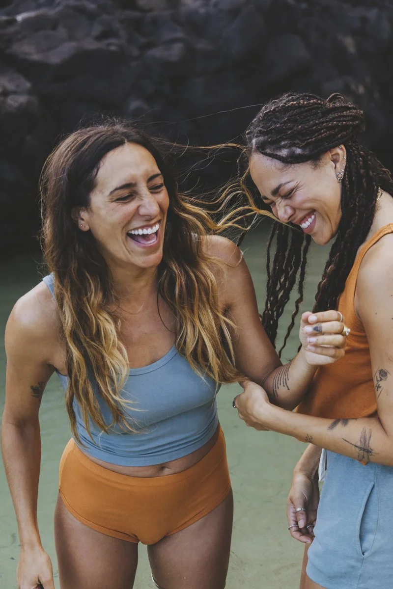 Two women laughing outdoors near rocks, wearing casual summer outfits.