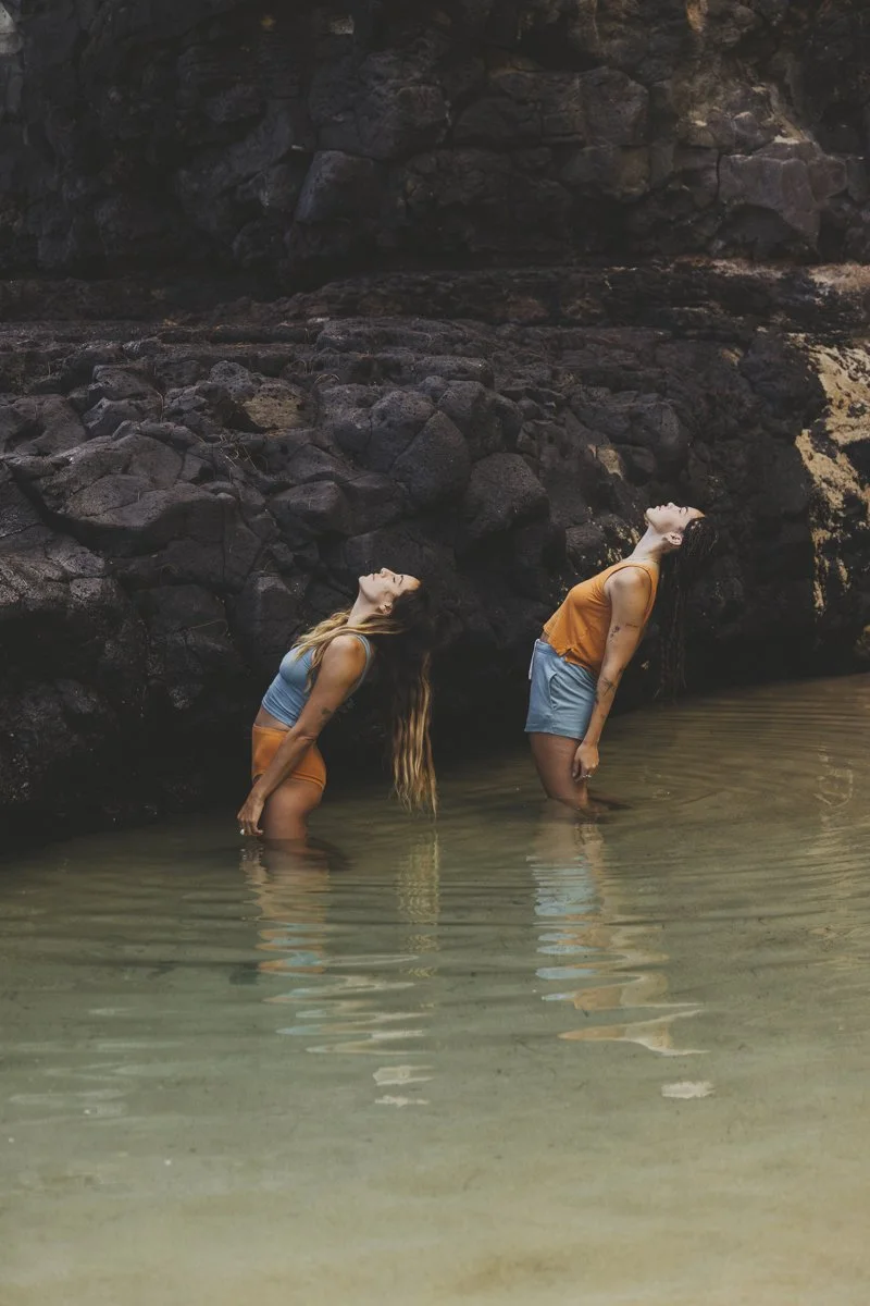 Two people standing in shallow water against a rocky backdrop, wearing swimwear, leaning back with heads tilted backwards.