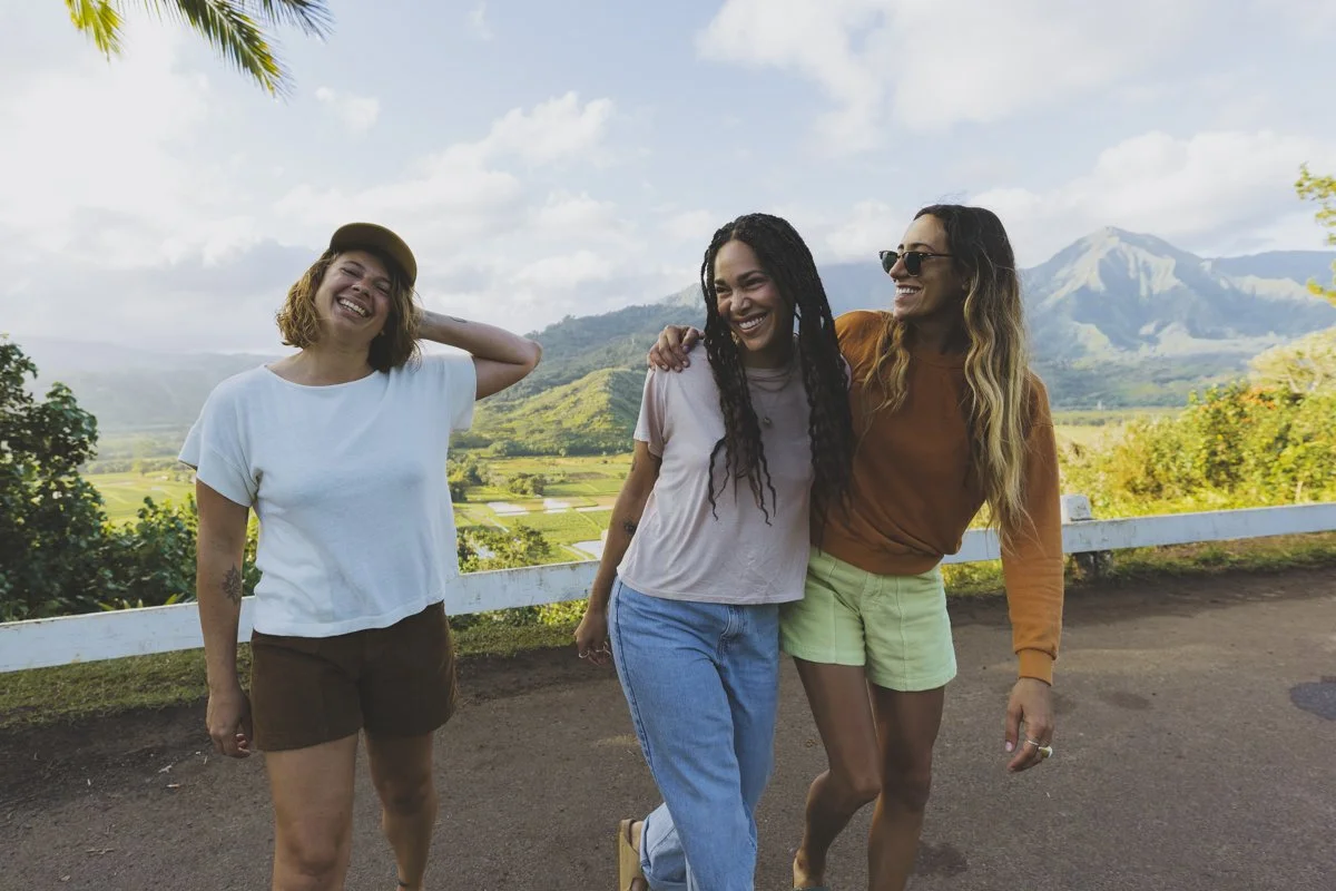 Three friends laughing and walking together outdoors in a mountainous and green landscape, with blue sky and clouds in the background.