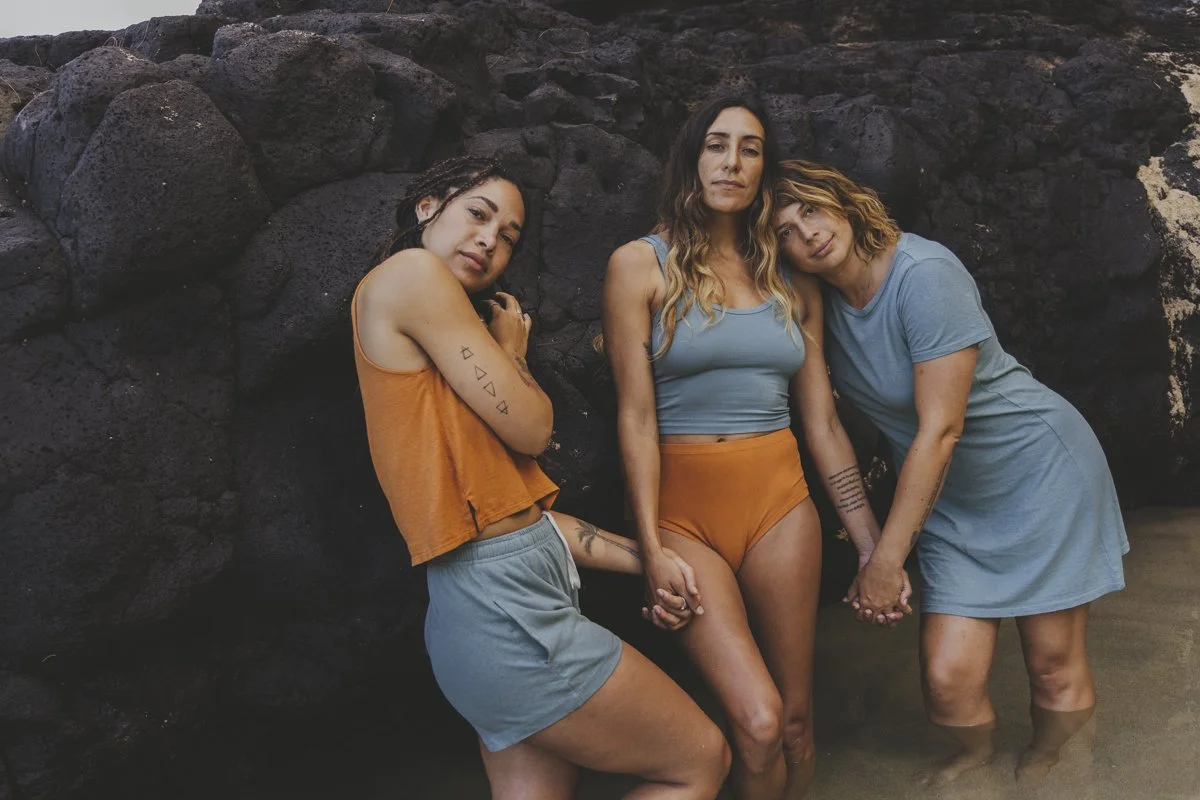 Three women standing on wet sand against black volcanic rocks, holding hands. They wear casual clothing in blue and orange tones, with tattoos visible on arms.