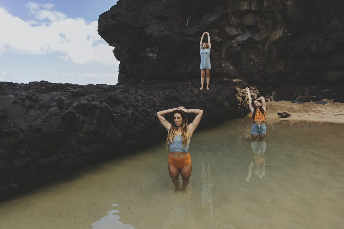 Three women in athletic clothing posing near rocky cliffs and shallow water on a beach.