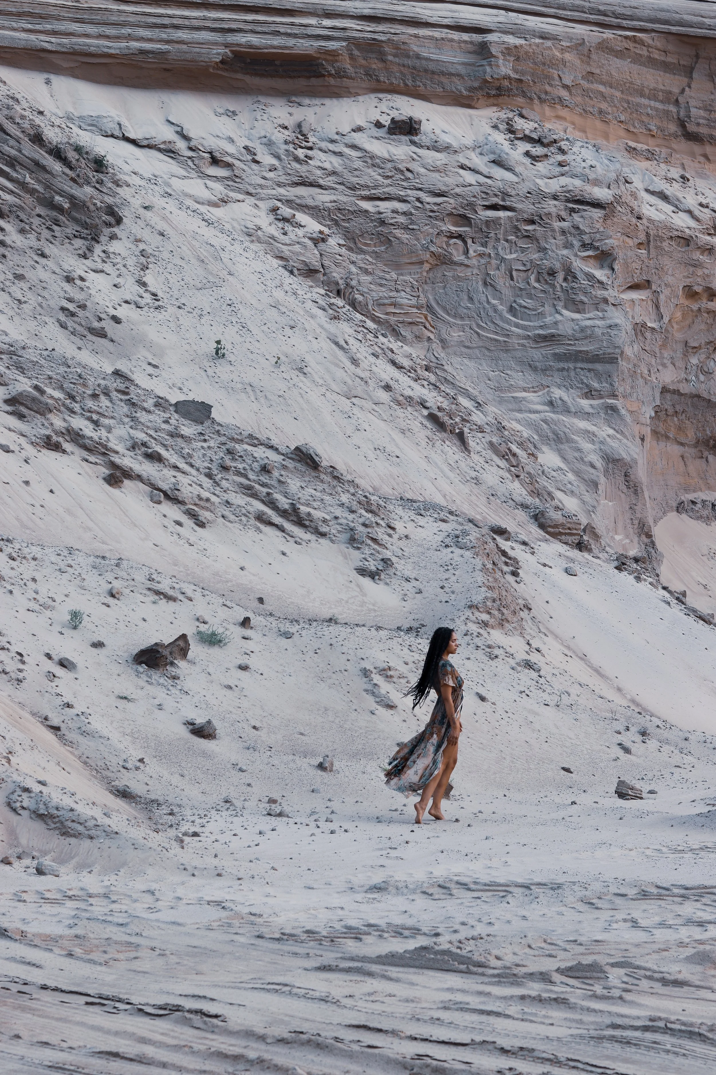 Woman in flowing dress standing on a rocky desert landscape with eroded cliffs.