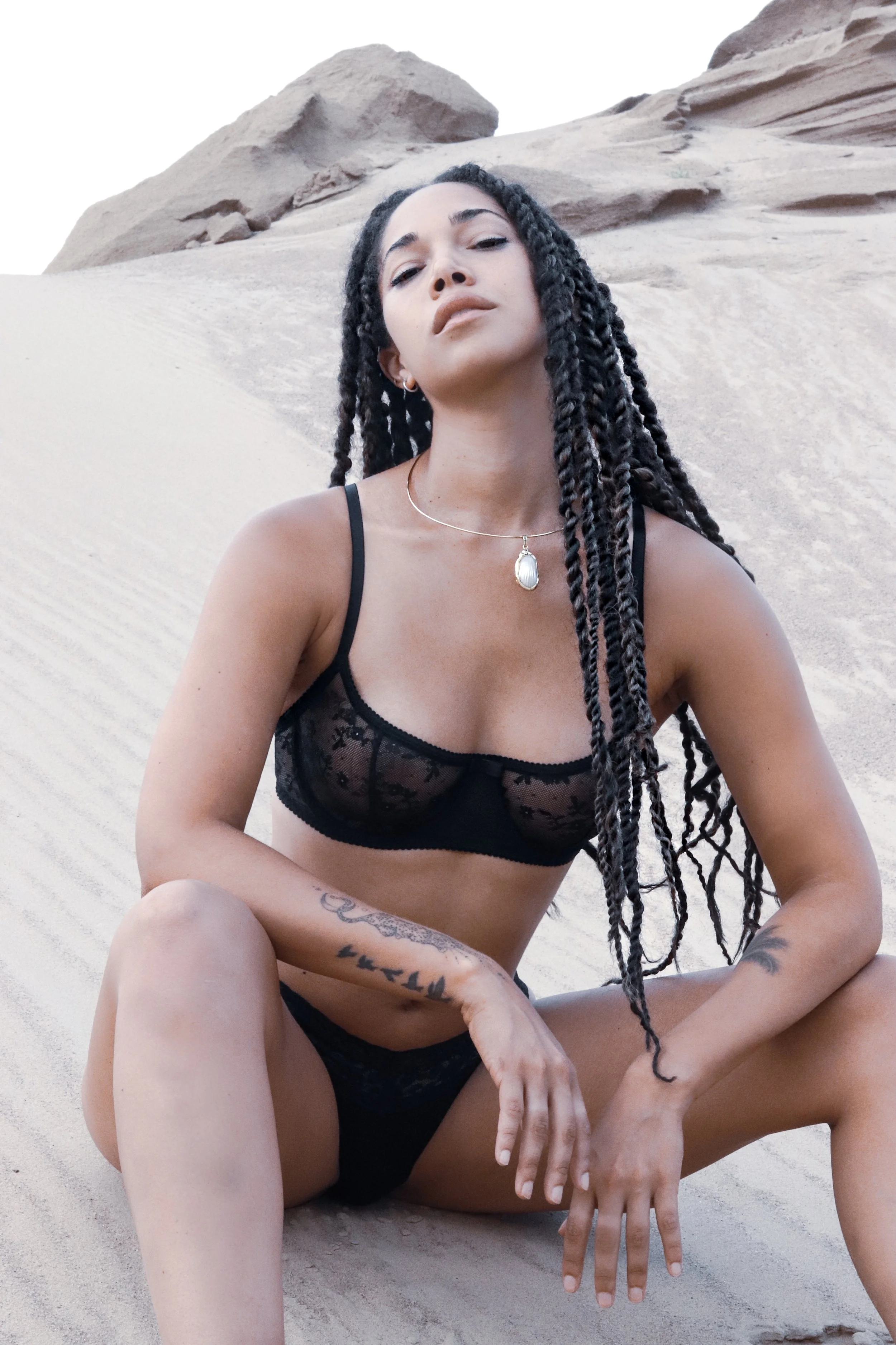 Person with braided hair wearing black lingerie sitting on sand
