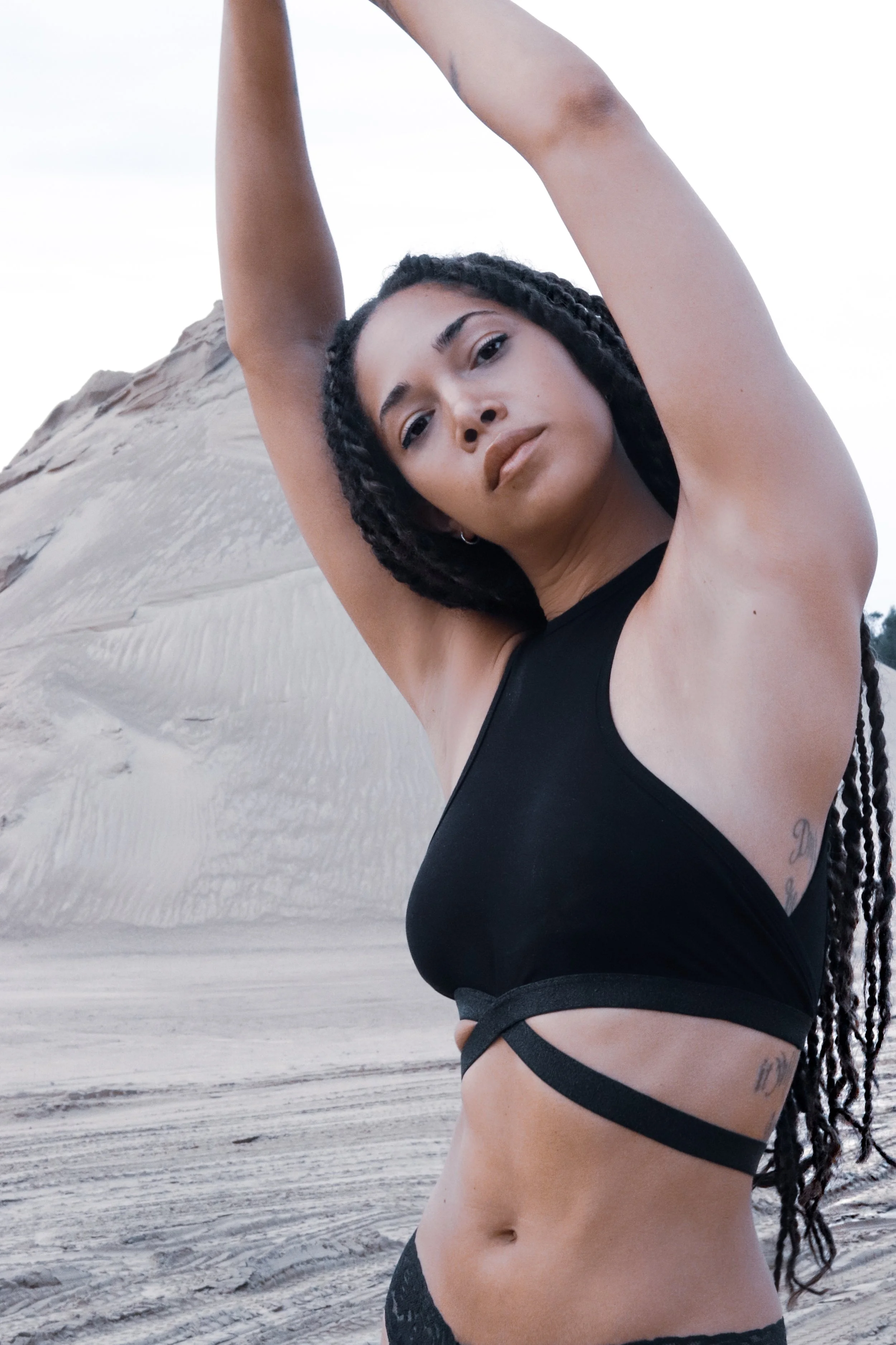 Woman with braided hair wearing black top, posing with arms raised in a desert landscape.