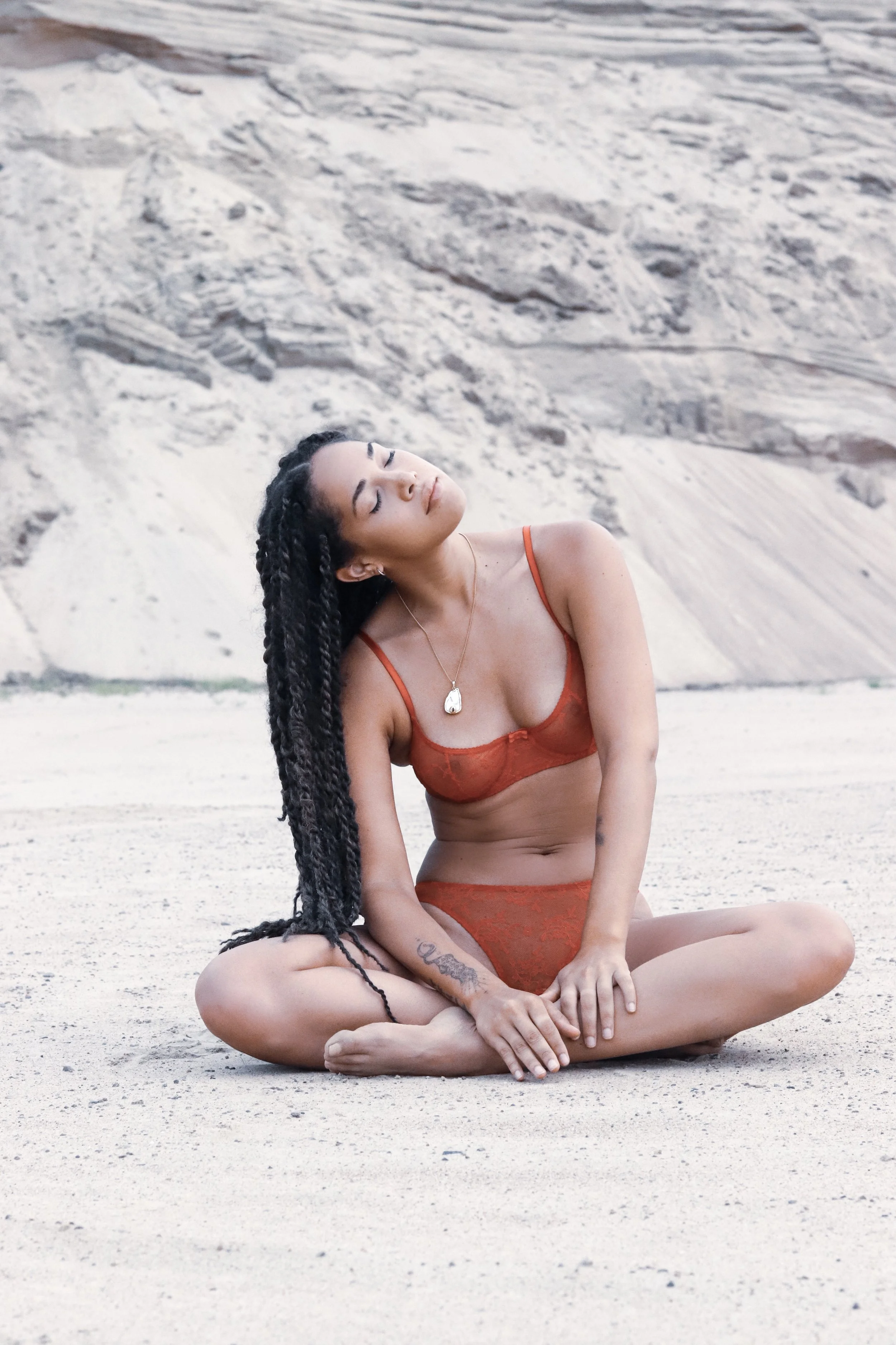 Woman in red lingerie sits cross-legged on a sandy terrain with rocky background.