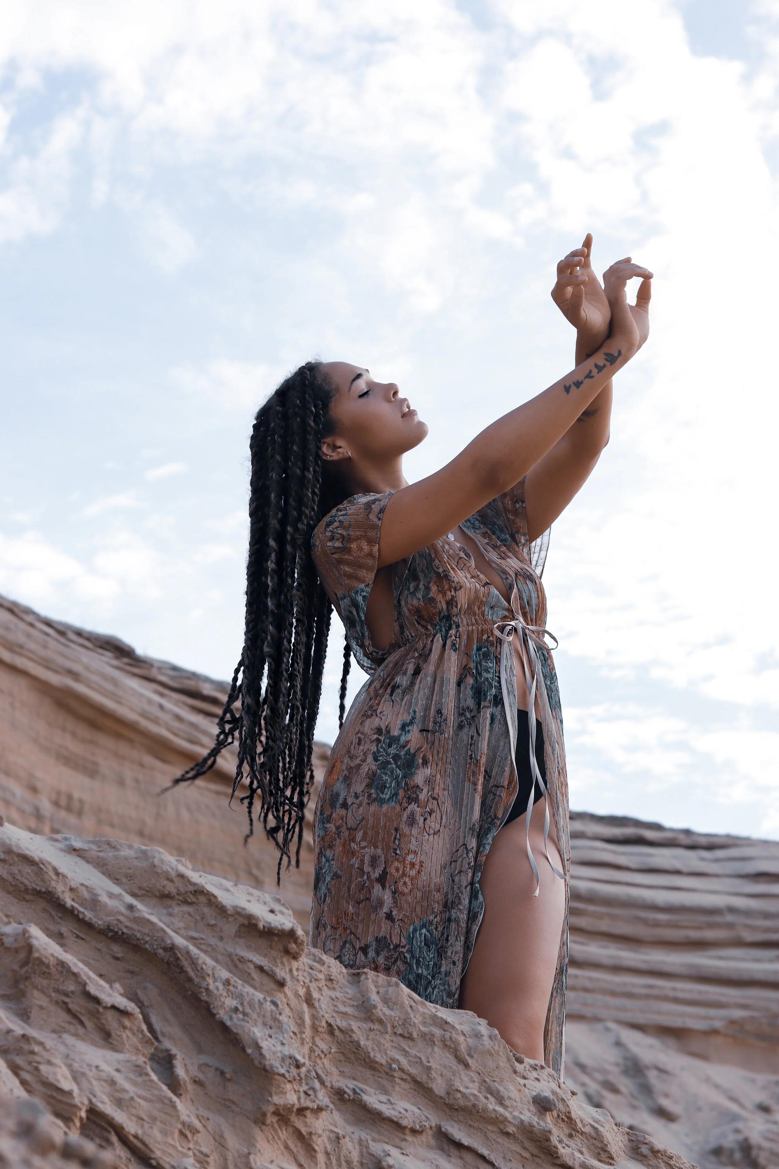 Woman with braids in a flowing dress posing against rocky scenery, arms raised.
