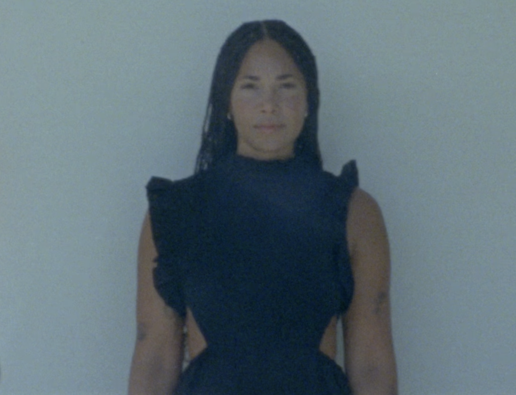 Woman in a dark sleeveless dress standing against a plain background.