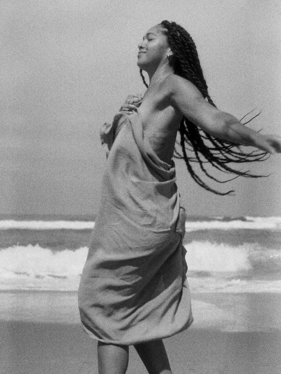Black and white photo of a person wrapped in a towel on a beach, with braided hair, facing sideways and arms outstretched, waves in the background.