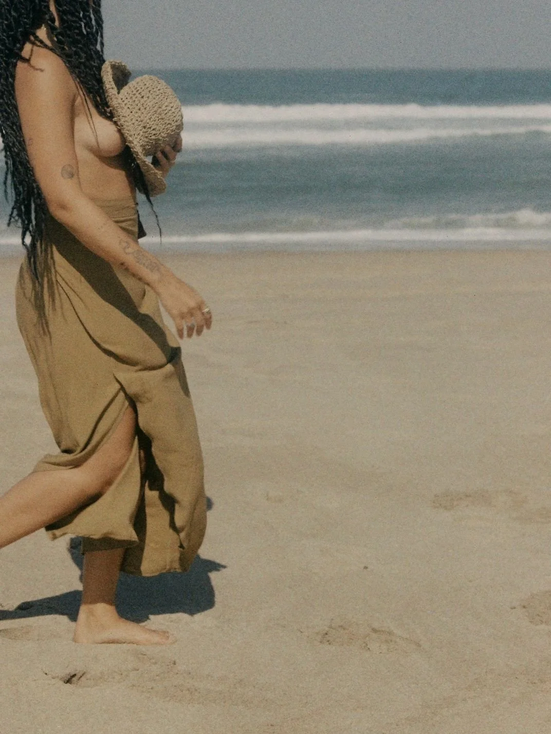 Person walking on a beach holding a straw hat and wearing a long skirt.