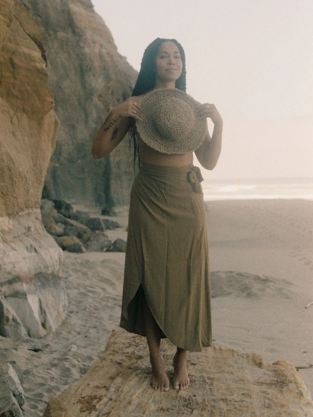 Woman standing on a rock at a beach holding a straw hat. She is wearing a brown skirt and there are cliffs in the background.