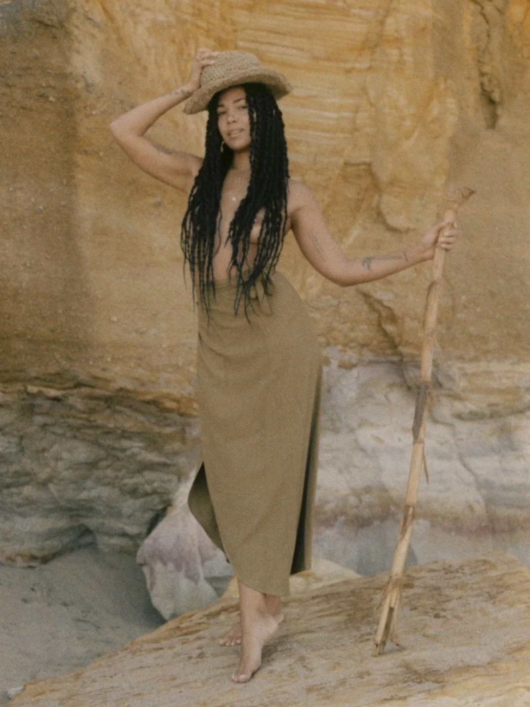 Person posing in front of a rocky background, wearing a wide-brimmed hat and a brown skirt, holding a walking stick.