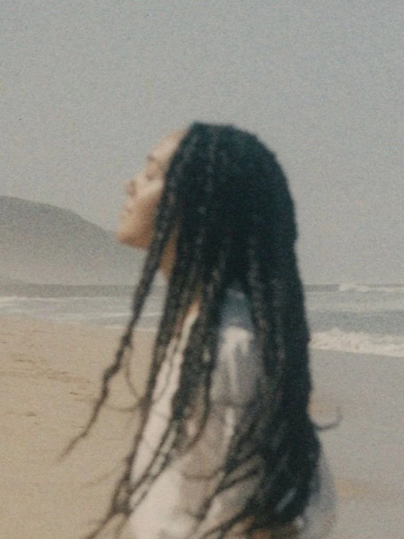 A person with long braided hair looking upwards on a beach, with waves in the background.