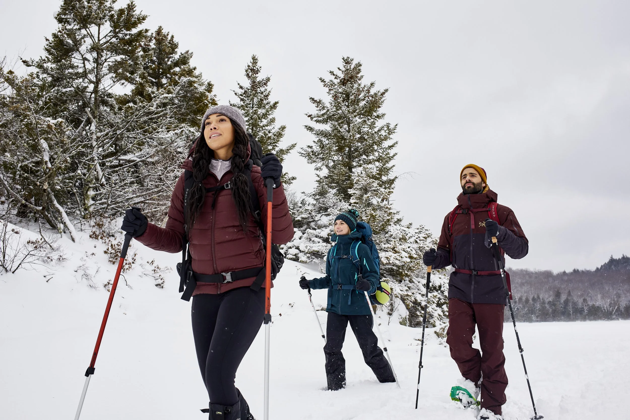 Three people wearing winter gear, snowshoeing in a snowy forest area, with trees in the background.