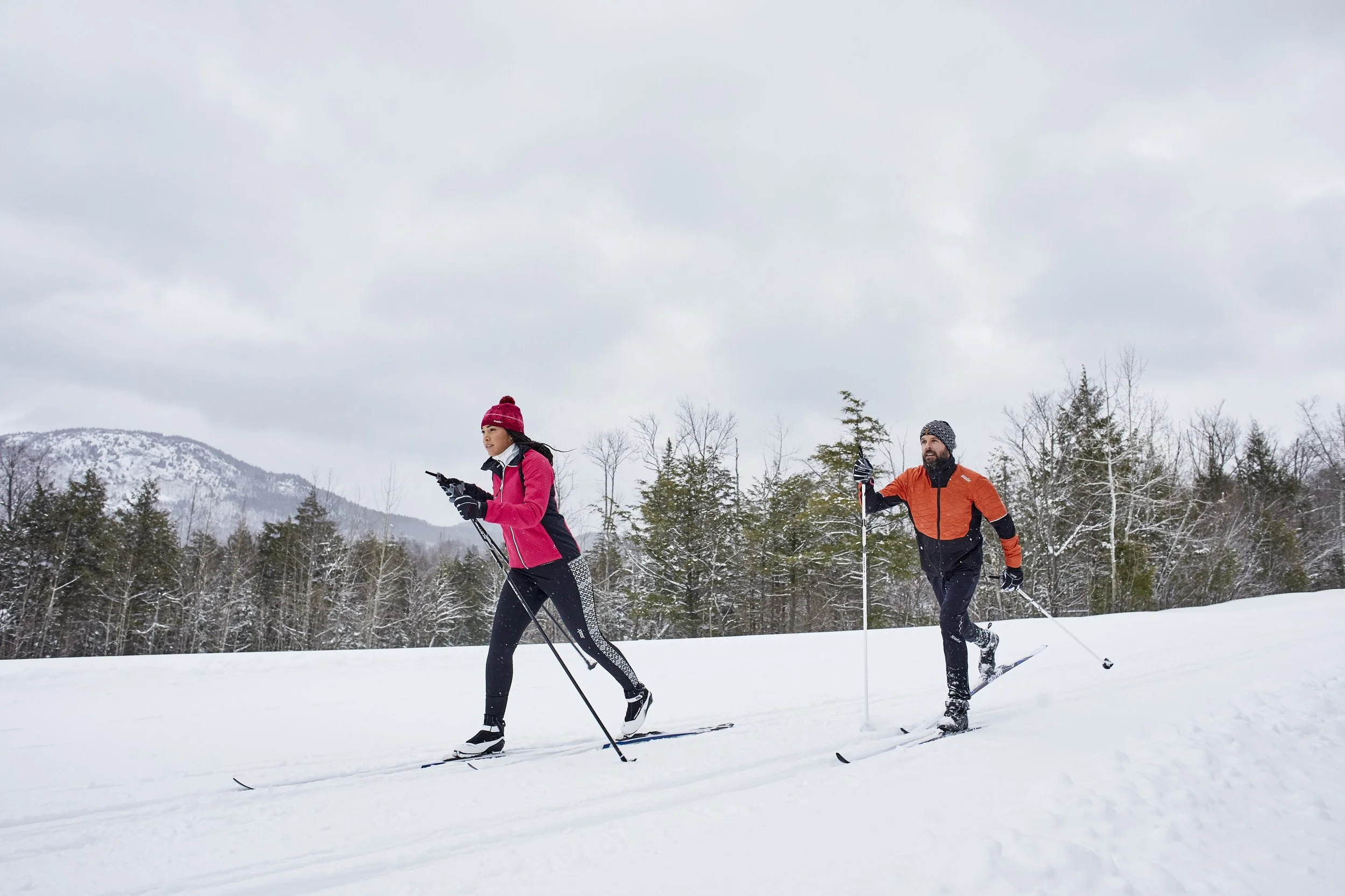 Two people skiing cross-country in a snowy landscape with trees and mountains in the background, both wearing colorful winter gear.
