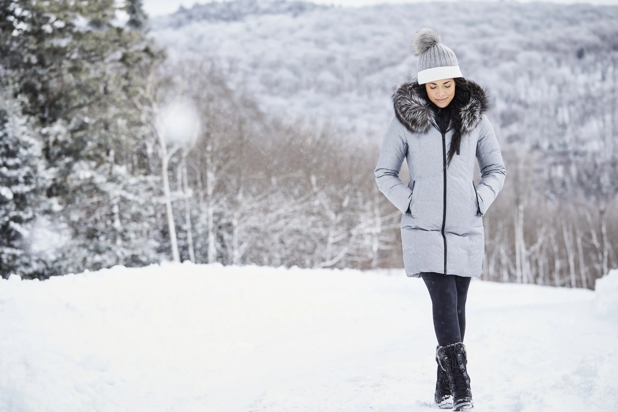 Woman in winter coat and beanie walking in snowy landscape.