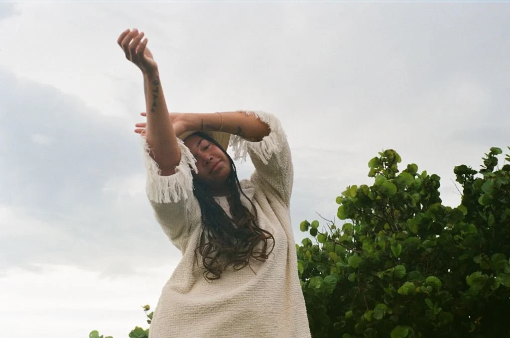 Person in a textured sweater with long hair, raising arms, standing outdoors in front of green foliage and a cloudy sky.