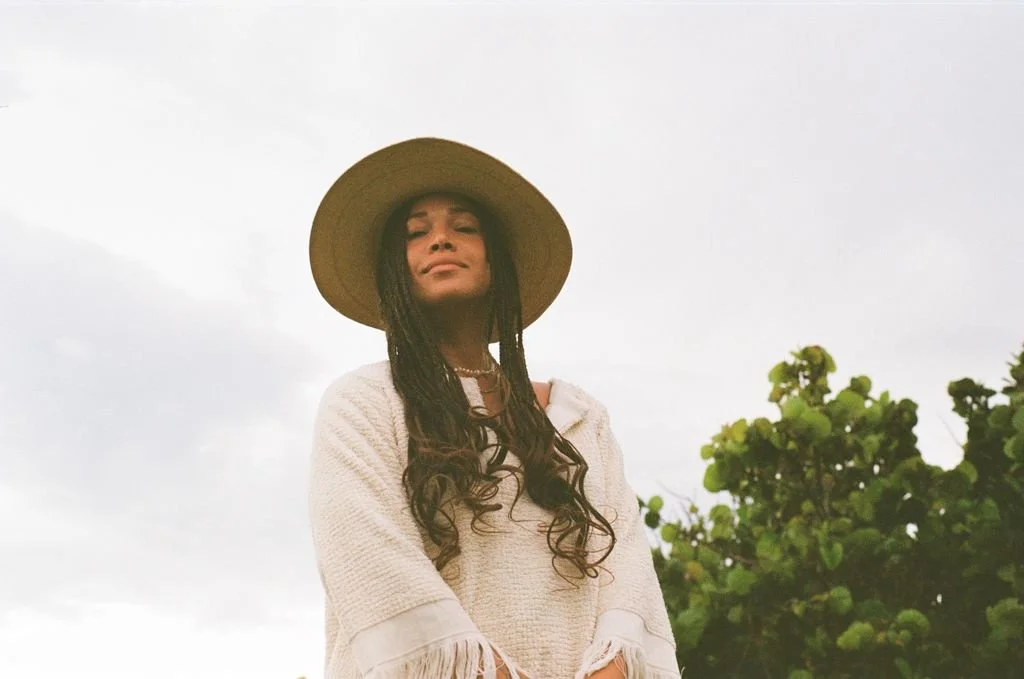 Woman with long hair wearing a wide-brimmed hat and a light-colored poncho standing outdoors with greenery and a cloudy sky in the background.