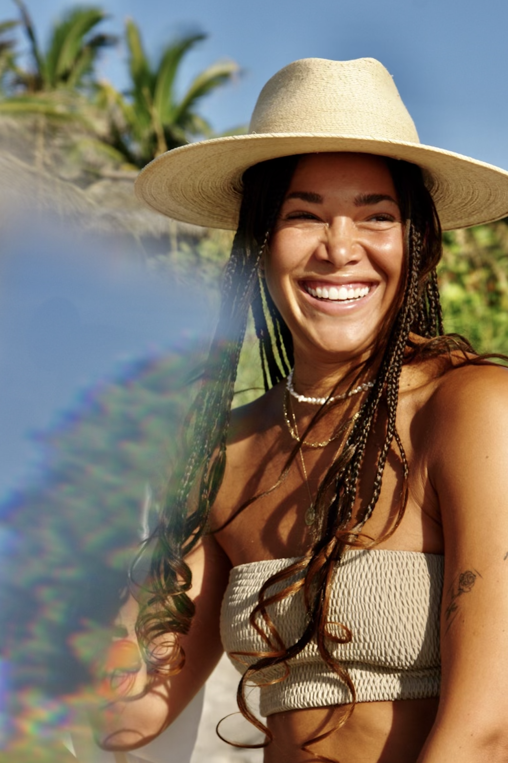 Smiling woman with long braids wearing a sun hat and strapless top outdoors.
