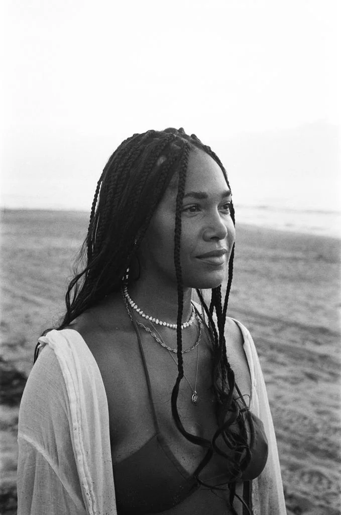 Black and white photo of a woman with braided hair standing on a beach, wearing a bikini top, a light cover-up, and layered necklaces.
