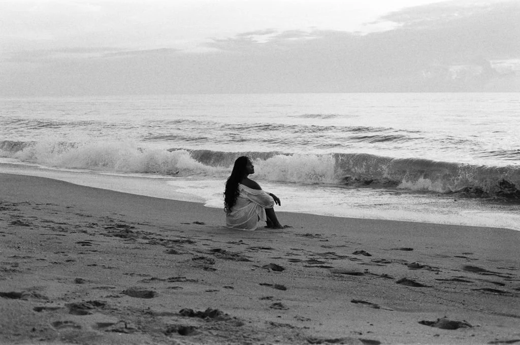 Black and white image of a person sitting on a sandy beach with waves in the background.