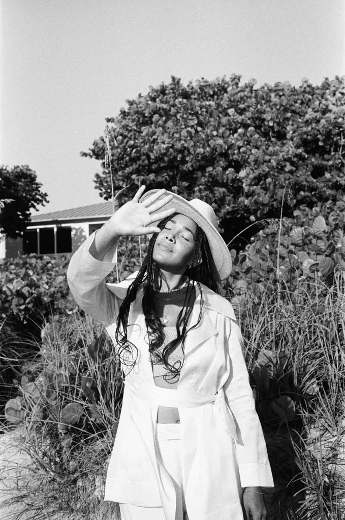 Black and white photo of a woman with long hair, wearing a hat and a light-colored outfit, standing outdoors with her hand raised, shielding her eyes from the sun. There are trees and a house in the background.