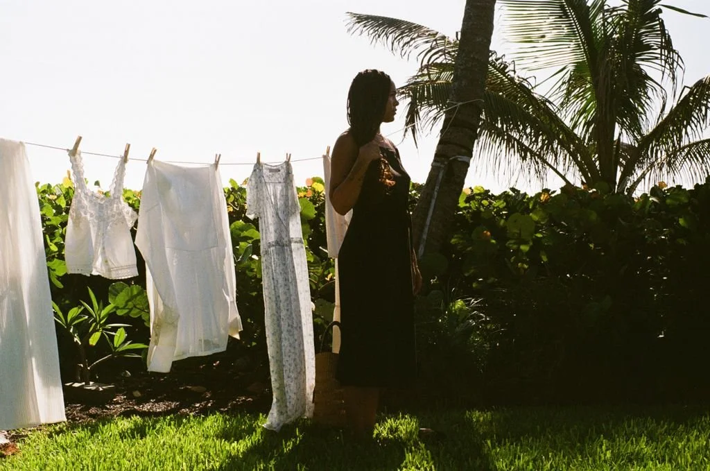Woman in a garden next to a clothesline with white garments, palm trees, and green foliage.