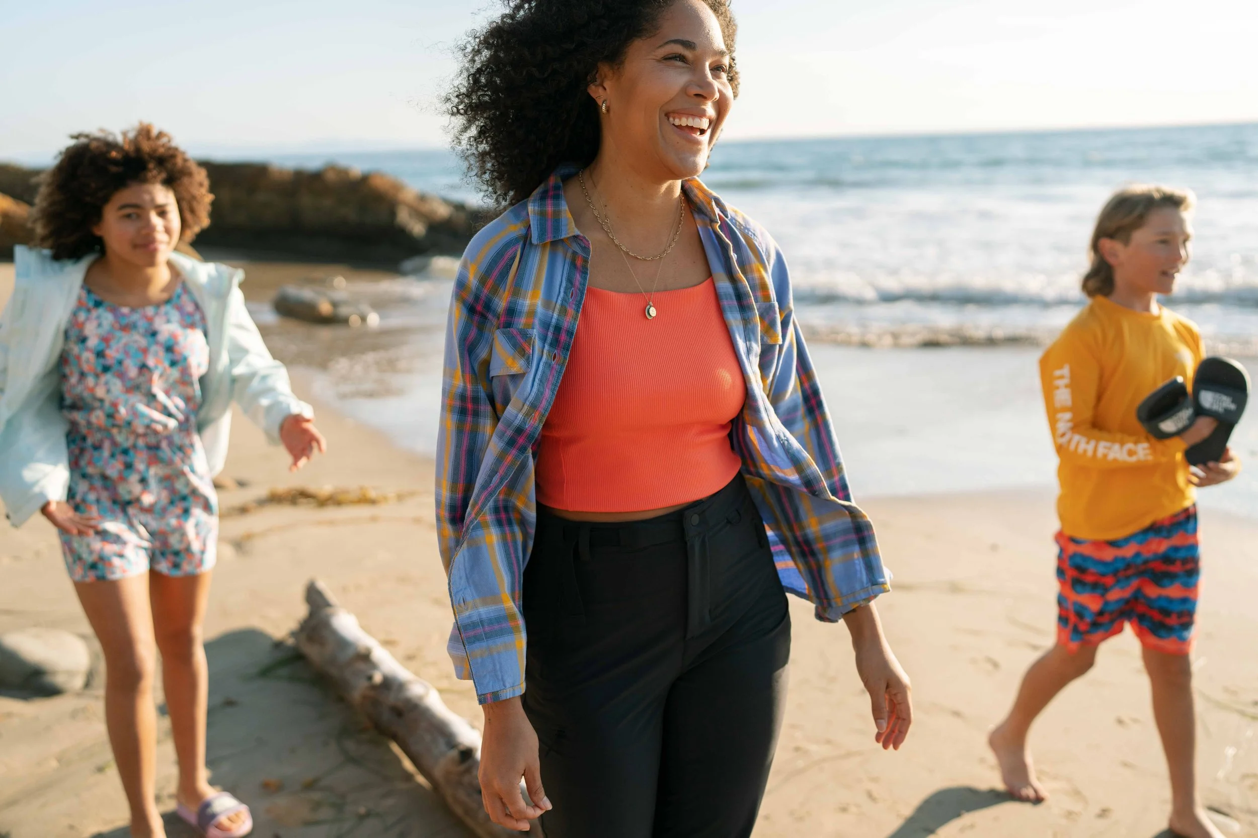Three people walking on a sunny beach, a smiling woman in the center wearing a plaid shirt and orange top, a child on the left in a floral outfit, and a child on the right in a yellow shirt and colorful shorts.