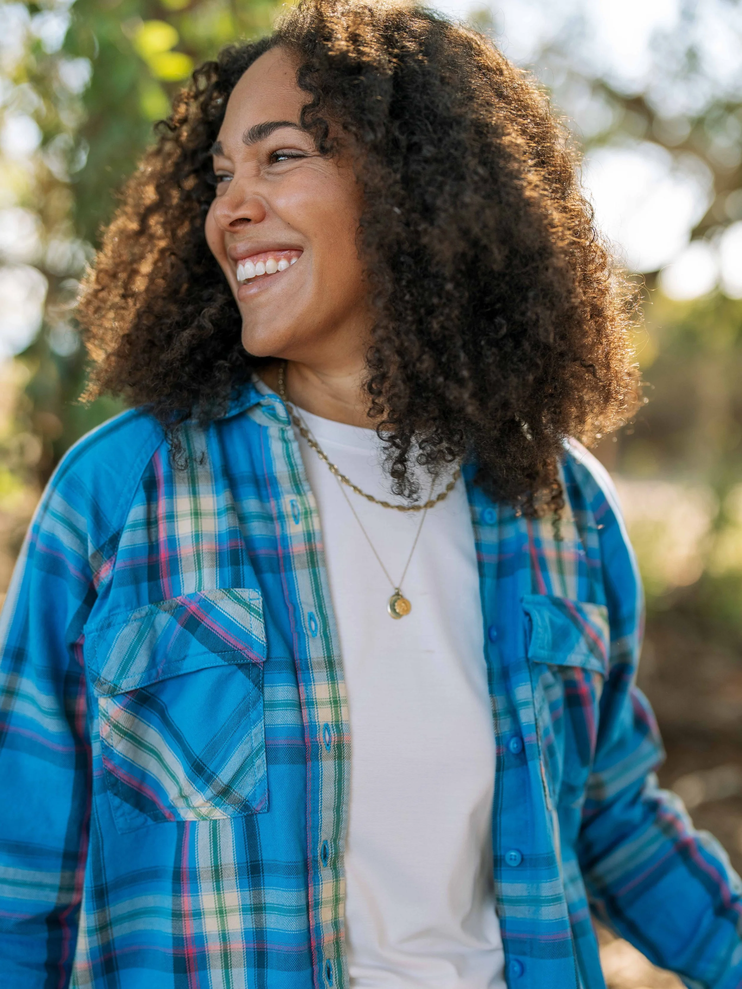 Person with curly hair smiling outdoors, wearing a blue plaid shirt over a white top.