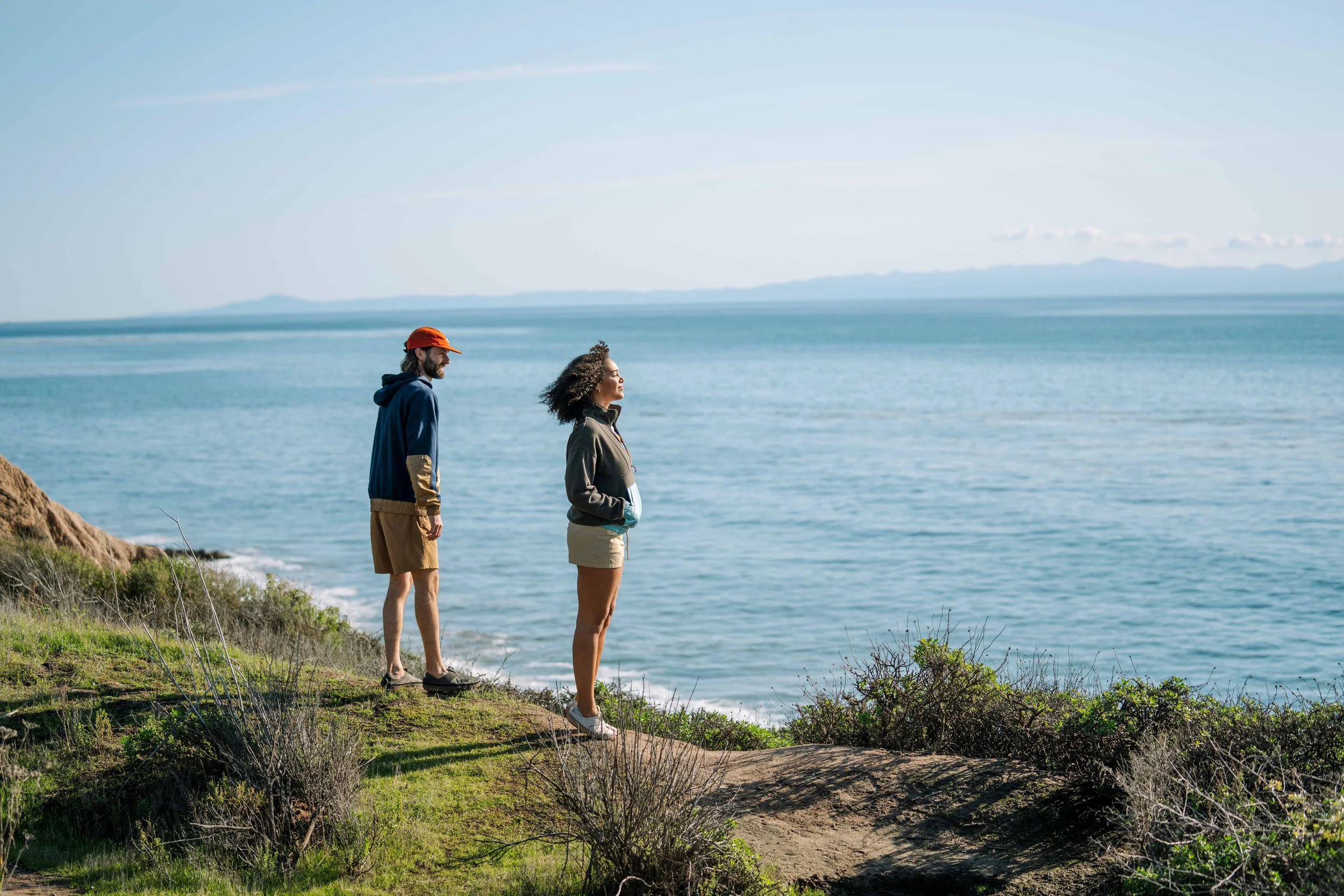 Two people standing on a grassy cliff overlooking the ocean, one wearing an orange hat and the other with curly hair, under a clear blue sky.