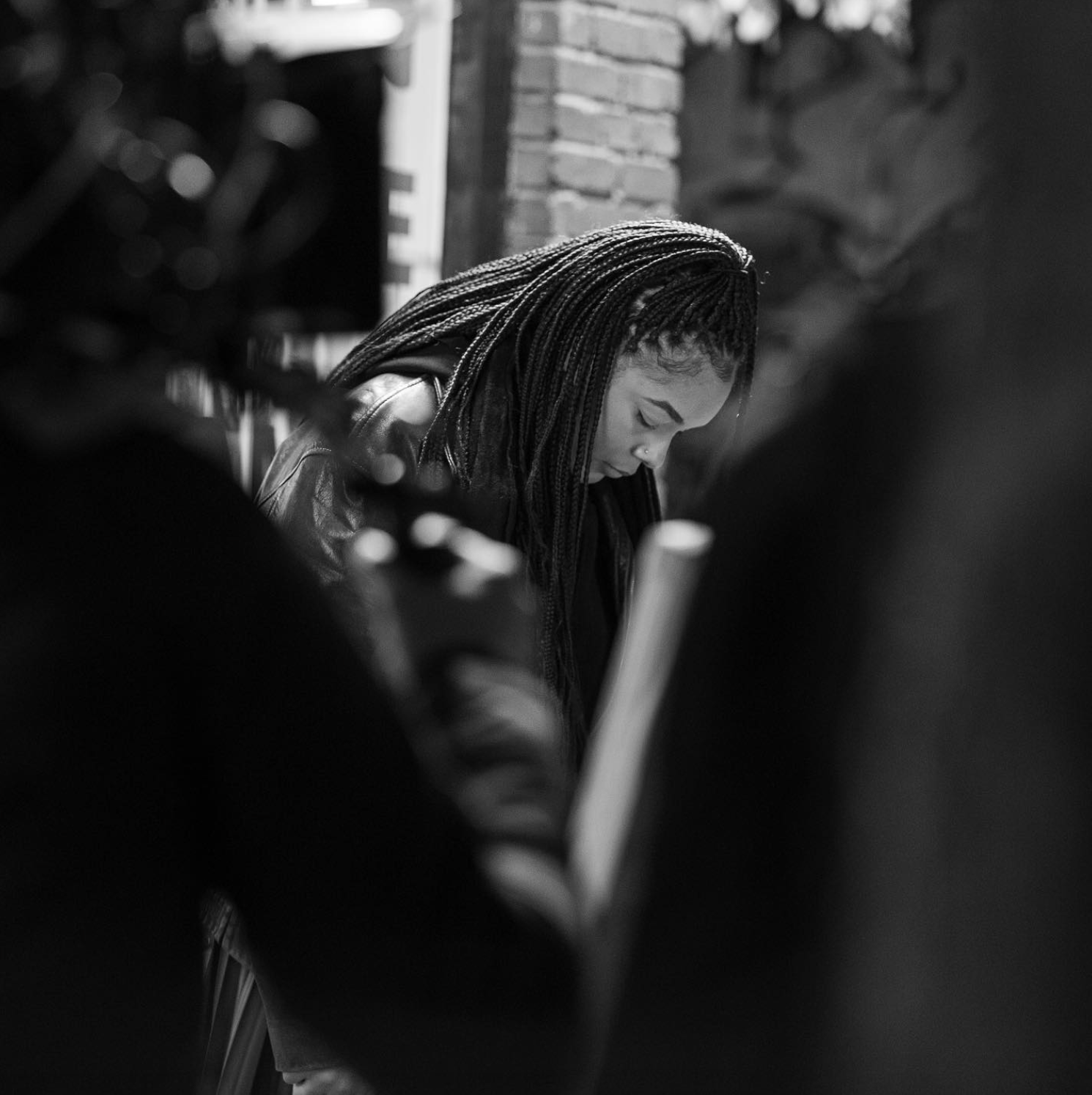 Black and white photo of a person with braided hair looking down in an outdoor setting, with blurred foreground and brick wall background.