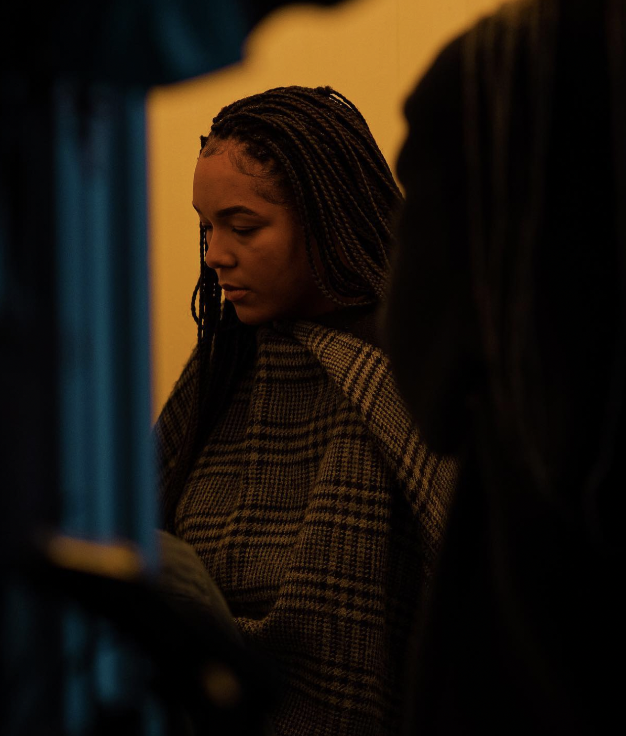 Person with braided hair wearing a checked plaid garment, looking down, in a dimly lit room.