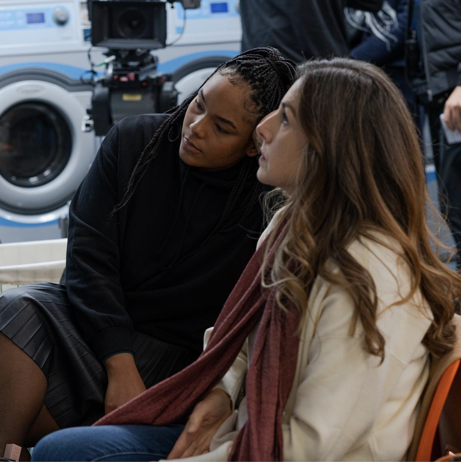 Two women sitting and talking in front of washing machines.