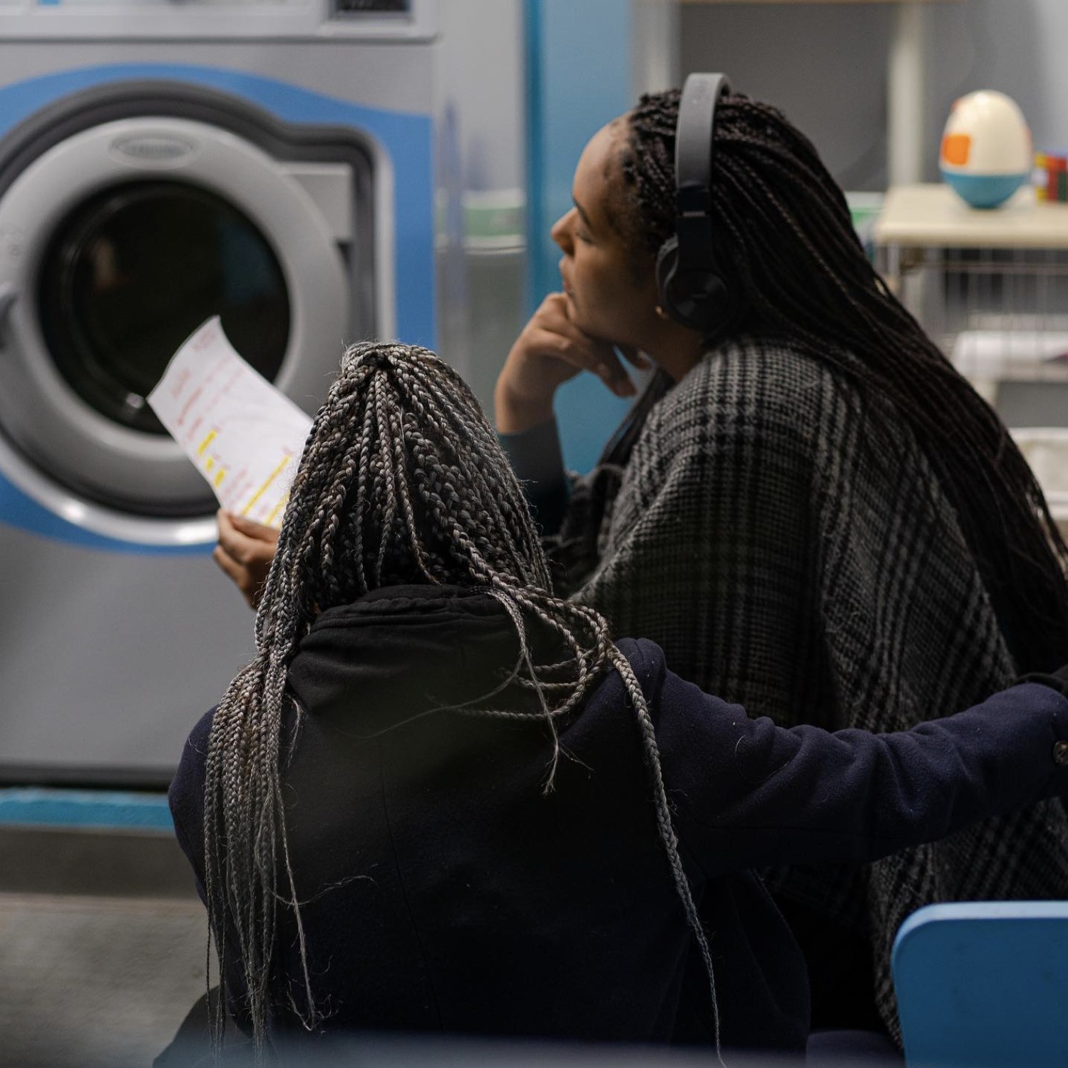 Two people in a laundromat sitting on blue chairs, one holding a piece of paper, with a washing machine in the background. Both have braided hair and one is wearing headphones.